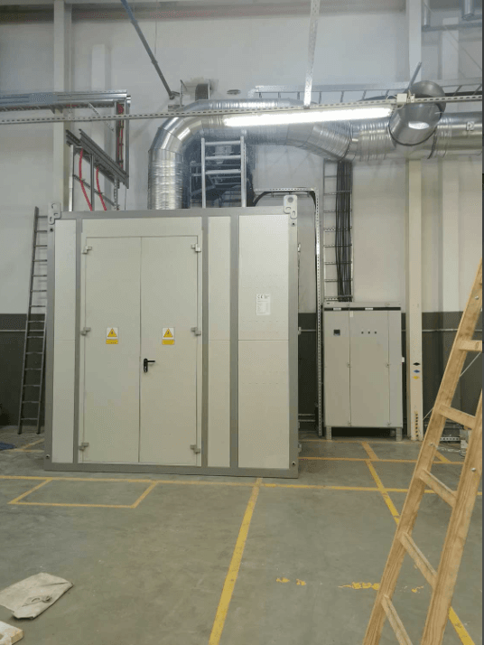 Zdjęcie zainstalowanej stacji transformatorowej wewnątrz hali, z widocznym skomplikowanym systemem wentylacji nad nią, szafą rozdzielczą obok i drabiną z boku, ilustrujące zakres prac instalacyjnych, co wiąże się z kosztem budowy stacji transformatorowej.